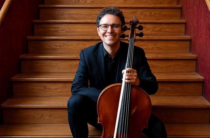 Malcolm KRAEGE, violoncelliste assis sur un escalier en bois tenant un violoncelle, souriant, en tenue de concert