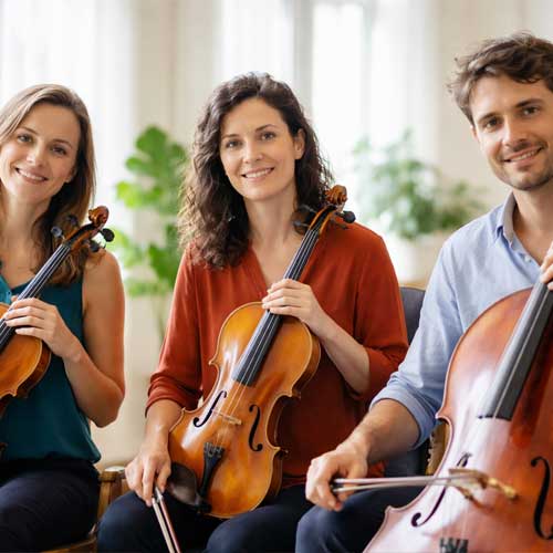 Trois musiciens classiques souriants — une violoniste, une altiste et un violoncelliste — assis côte à côte avec leurs instruments dans un espace lumineux.